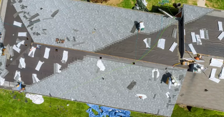 A roof in the process of being replaced with bundles of shingles on the roof, some underlayment exposed and workers adding new shingles.