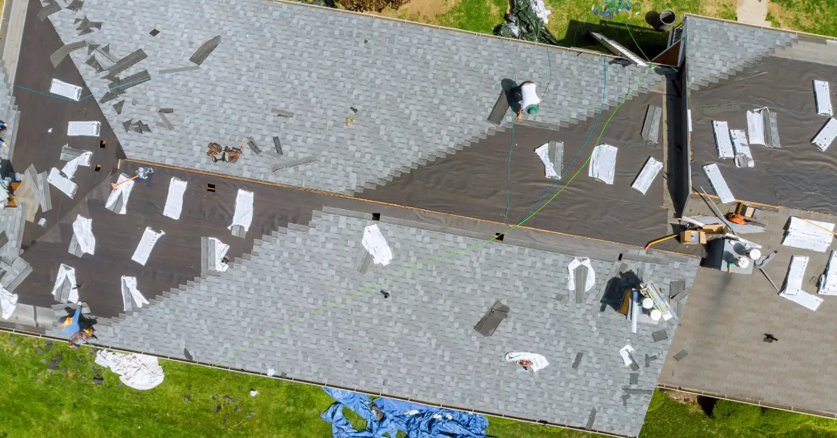 A roof in the process of being replaced with bundles of shingles on the roof, some underlayment exposed and workers adding new shingles.
